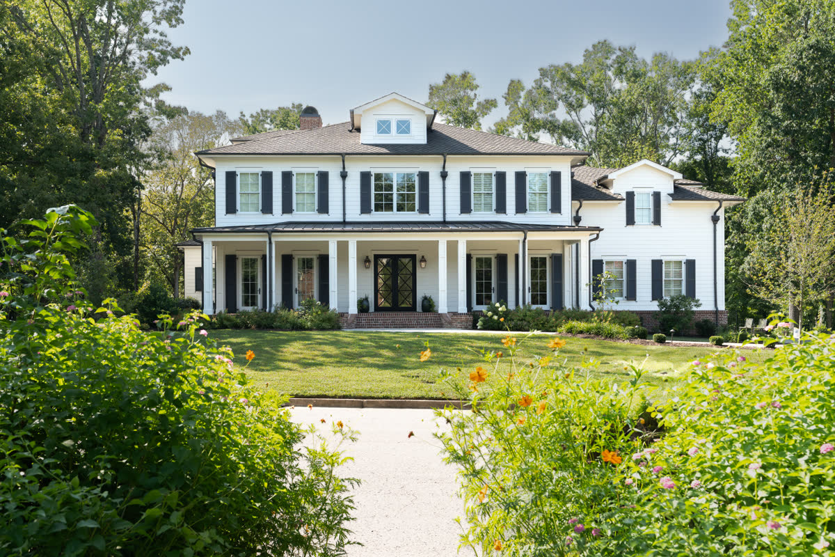 A custom-built white home with black shutters and a covered front porch, set behind a lawn and flower garden.